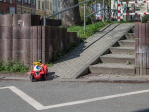 Das Bild ist ein Foto eines Zugangs zu dem Spielplatz Fichteplatz in Jena. Er besteht aus einer steilen Rampe und einer Treppe. Die Rampe geht abschüssig auf eine Straße zu, auf der Straße ist durch eine Zickzack Linie ein Halteverbot eingezcihent. Ein Bobbycar steht am Ende der Rampe.